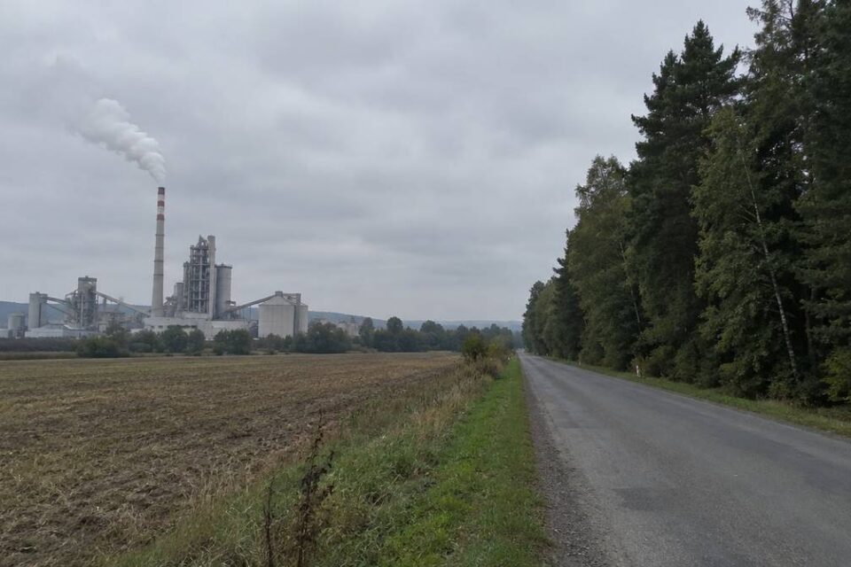 Cementownia z dymiącym kominem i las mieszany po drugiej stronie pola, pod zachmurzonym niebem