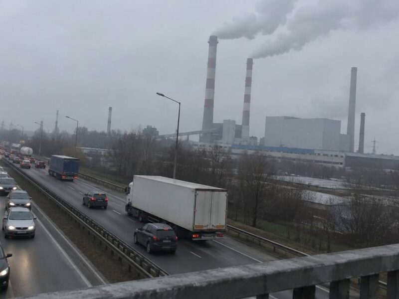 Ruchliwa droga z samochodami i ciężarówkami, w tle kominy zakładu przemysłowego, z których unosi się dym