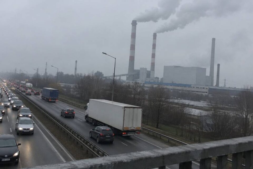 Ruchliwa droga z samochodami i ciężarówkami, w tle kominy zakładu przemysłowego, z których unosi się dym
