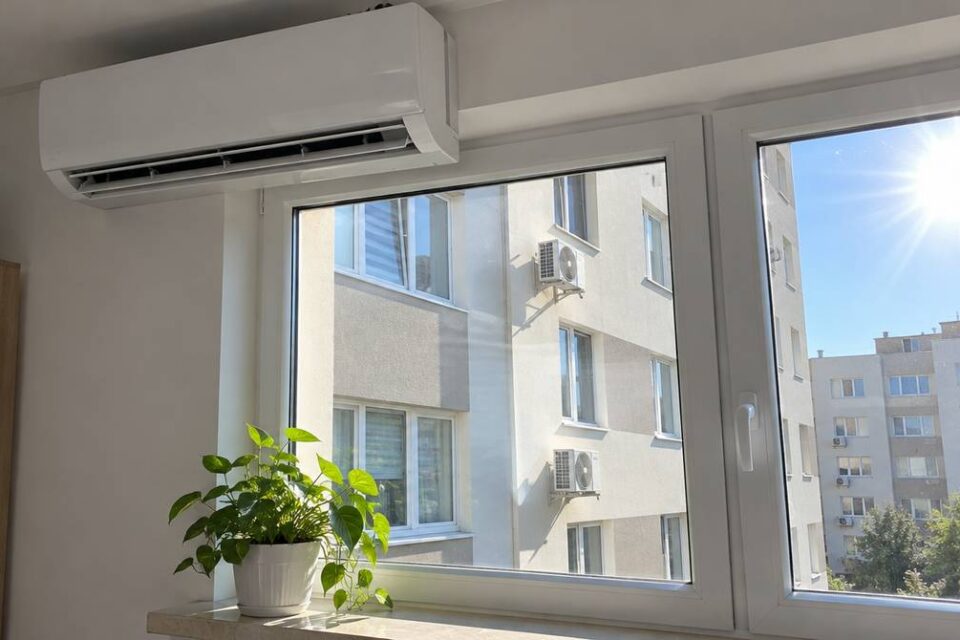 Jednostka wewnętrzna klimatyzatora split nad oknem, za szybą blok z jednostkami zewnętrznymi w upalny dzień