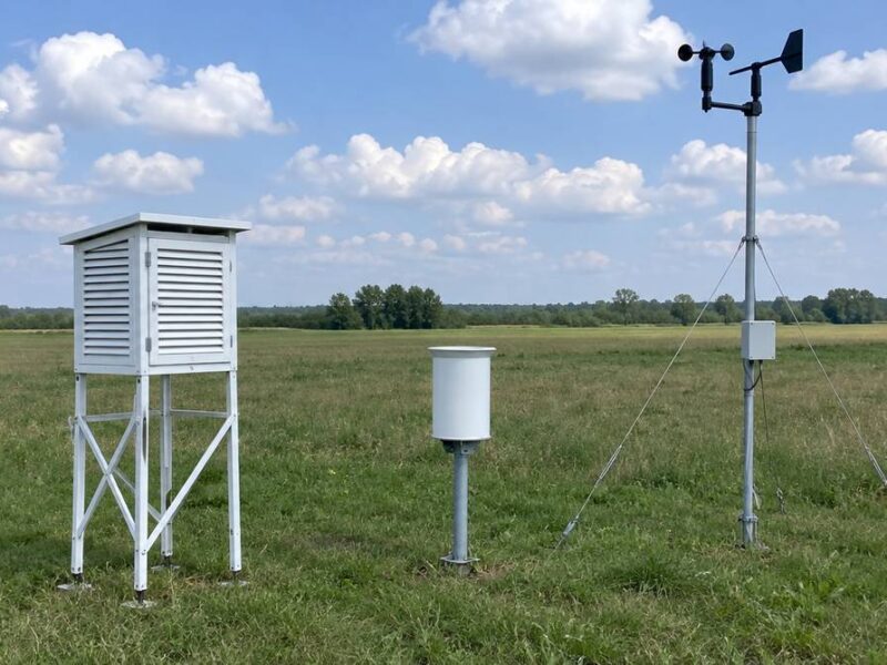 Stacja meteorologiczna na łące z białą klatką meteorologiczną, deszczomierzem i masztem z wiatromierzem